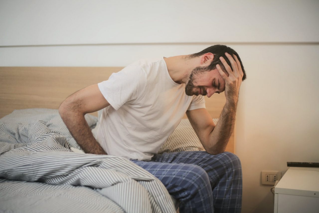 A man holding his forehead on the edge of his bed.