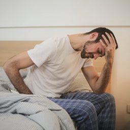 A man holding his forehead on the edge of his bed.