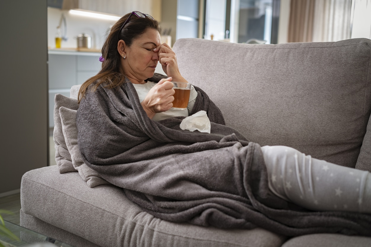 Sick woman resting on sofa drinking hot tea and massaging her nose.